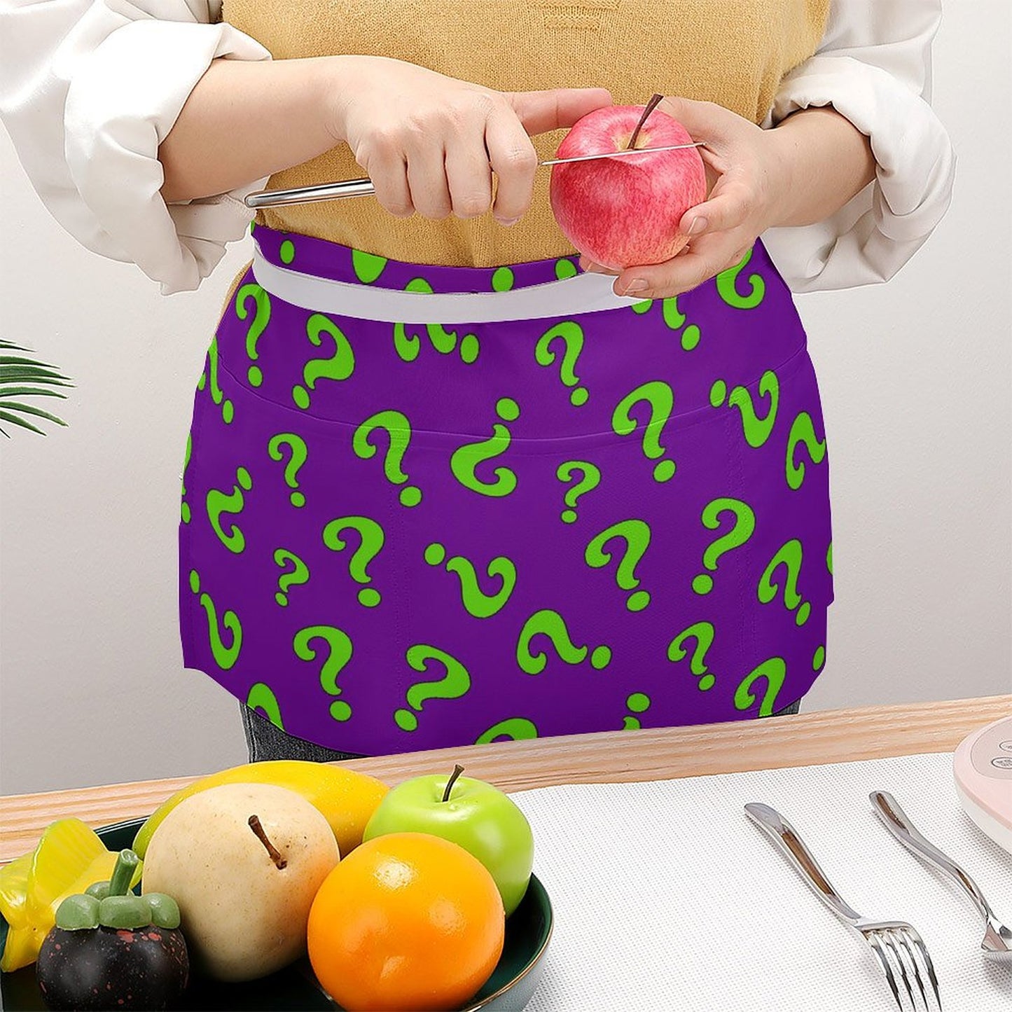 Person wearing a purple apron with green question marks, holding an apple, standing behind a table with fruits and utensils.